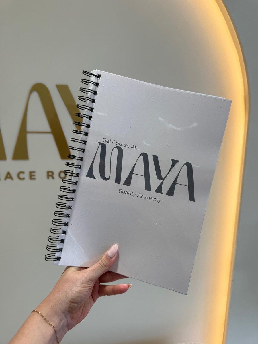 A Maya Beauty Academy gel course workbook held up in front of the MAYA arch at the salon