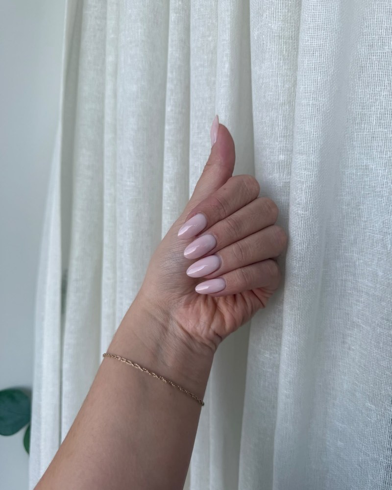 Soft pink almond nails with a glossy finish, hand resting against a white curtain